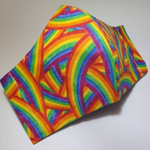 May include: A rainbow-colored face mask with a pattern of overlapping rainbow arches. The mask is made of fabric and has a three-dimensional shape.