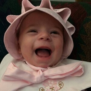 May include: A smiling baby wearing a pink bonnet and bib. The bonnet has a scalloped brim, and the bib features a large bow and embroidered floral design. The baby's mouth is wide open, showing their tongue.