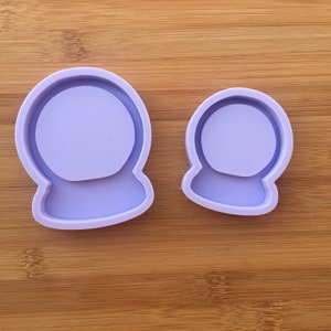 Könnte beinhalten: Zwei hellviolette Silikonformen in Form einer Schneekugel. Die Formen bestehen aus einem kreisförmigen oberen Teil und einem breiteren, rechteckigen Sockel. Das Silikonmaterial ist glatt und undurchsichtig mit einem erhöhten Rand um die Vertiefung.