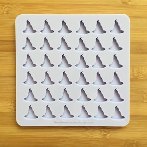 May include: White silicone mould with 48 individual cavities shaped like witch hats. The mould is on a wooden surface. The text "thecraftsandglittereshop.com" is at the bottom of the mould.