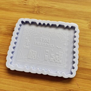 May include: A light purple silicone mold with a scalloped edge. The mold features a Halloween-themed design with the text "DNE CEMETERY" and illustrations of a tombstone, cobwebs, and a bat. The design is embossed within the square mold.