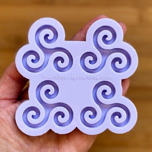 Op de afbeelding: Een lichtpaarse siliconen mal met een Keltisch triskelion ontwerp. De mal heeft een wolkvorm met zes triskelions, elk met drie in elkaar grijpende spiralen. De tekst "thecraftsandglittershop.com" is op de mal gedrukt.