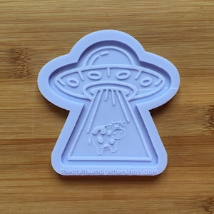 Könnte beinhalten: Lilafarbene Silikonform eines UFOs, das eine Kuh entführt. Die Form ist dazu bestimmt, einen Anhänger aus Harz oder Polymer Clay zu erstellen. Der Text "thecraftsandglittershop.com" befindet sich am unteren Rand der Form.