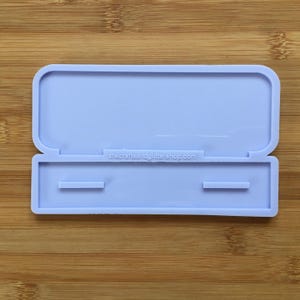 Puede incluir: Un molde de silicona azul claro con forma de caja rectangular pequeña con tapa con bisagras. El molde tiene dos hendiduras rectangulares en la sección inferior. El texto "thecraftsandglittershop.com" está impreso en el molde.
