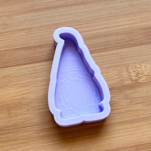 Pode incluir: Um molde de silicone lavanda em forma de gnomo. O molde tem um design detalhado do chapéu, rosto e barba do gnomo. O molde está sobre uma superfície de madeira.