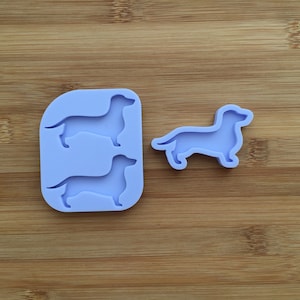 Pode incluir: Um molde de silicone roxo claro com duas silhuetas de cão salsicha em baixo-relevo e uma peça de silicone separada a condizer em forma de cão salsicha. O molde tem cantos arredondados e um acabamento mate liso, concebido para artesanato ou culinária.