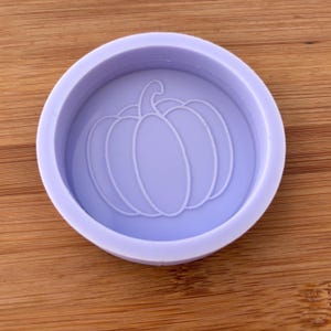 May include: A pale purple, circular mould featuring a pumpkin design. The pumpkin is outlined in white and is centrally positioned within the mould. The mould is on a wooden surface.