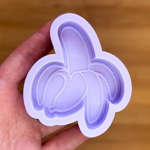 Puede incluir: Un molde en forma de plátano de color lila claro, con un diseño de plátano pelado. El molde tiene una construcción en capas, con la forma del plátano claramente definida. El diseño general es suave y limpio, adecuado para proyectos de manualidades o repostería.