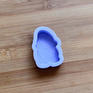 Puede incluir: Un molde de silicona de color púrpura claro con forma de gnomo. El molde presenta un contorno detallado del sombrero, la barba y la ropa del gnomo. El molde está sobre una superficie de madera.