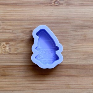 Puede incluir: Un molde de silicona lavanda con forma de gnomo. El gnomo tiene un gorro puntiagudo, una barba y un cuello decorativo. El molde está sobre una superficie de madera.