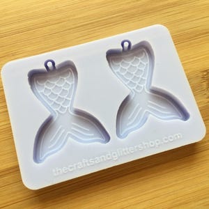 Puede incluir: Un molde de silicona blanco con dos formas de cola de sirena. Cada cola tiene un agujero en la parte superior para colgar. El molde está etiquetado con "thecraftsandglittershop.com".