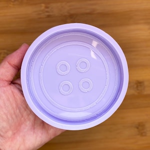 May include: A light purple, circular mould shaped like a large button. The mould has four small holes in the centre and concentric circles around the edge, creating a detailed button design. The mould is held in a hand.
