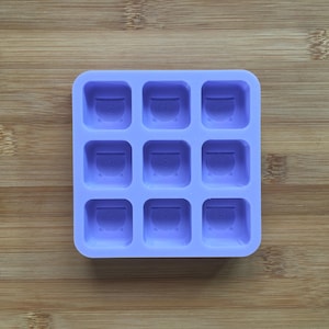 May include: A lavender silicone ice cube tray with nine square molds. Each mold features a cauldron design. The tray is made of flexible silicone and is designed for easy ice removal. The tray is on a wooden surface.