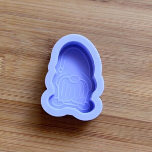 Puede incluir: Un molde de silicona lavanda con forma de cara de gnomo. El molde presenta un gorro puntiagudo, una nariz redonda y una larga barba. El molde está diseñado para manualidades y se coloca sobre un fondo de madera.