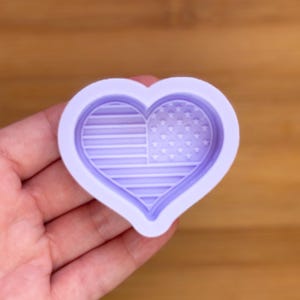 Puede incluir: Un molde en forma de corazón de color lila claro con un diseño de la bandera estadounidense. El interior presenta el patrón de estrellas y rayas en detalle. El borde exterior del molde es liso y redondeado, ideal para manualidades patrióticas.