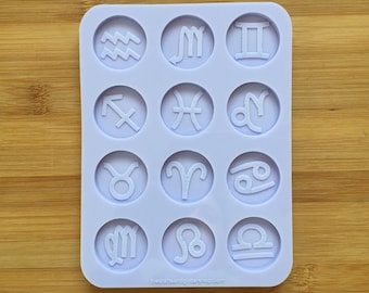 Könnte beinhalten: Eine hellviolette, rechteckige Silikonform mit zwölf kreisförmigen Vertiefungen, die jeweils ein erhabenes Sternzeichen-Symbol enthalten. Der Text thecraftsandglittershop.com ist unten in der Mitte der Form mit abgerundeten Ecken eingeprägt.