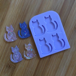 Puede incluir: Un molde de silicona con cuatro cavidades en forma de gato. Se muestran cuatro amuletos de gato de resina brillante junto al molde.