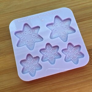 Puede incluir: Un molde de silicona lila con cinco cavidades en forma de copo de nieve. El molde tiene el texto "thecraftsandglittershop.com" impreso. Los copos de nieve tienen diseños detallados y delicados, perfectos para proyectos de manualidades.