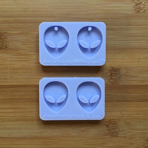 Puede incluir: Molde de silicona de dos partes para hacer pendientes con forma de cabeza de extraterrestre. El molde es de color morado claro y tiene dos cavidades, cada una con forma de cabeza de extraterrestre con dos ojos y un pequeño agujero en la parte superior para colgar.