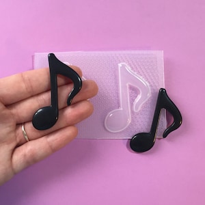 Può includere: Uno stampo in silicone trasparente con due forme di note musicali nere. Lo stampo è su una superficie rosa.