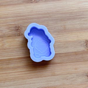 Puede incluir: Un molde de silicona de color lila claro con forma de gnomo. El molde tiene un sombrero, barba y detalles del cuerpo. El molde está sobre una superficie de madera.