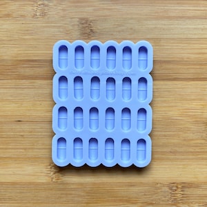 Può includere: Uno stampo in silicone di colore lilla chiaro con 24 cavità individuali a forma di pillole. Lo stampo è su una superficie di legno. Thecraftsandglittersshop.com è stampato sullo stampo.