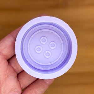 Può includere: Uno stampo circolare lilla chiaro per la creazione artigianale, con un design a bottone. Lo stampo presenta anelli concentrici e quattro piccoli fori equidistanti al centro, creando la forma di un bottone. Lo stampo è tenuto in una mano.