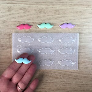 Può includere: Uno stampo in plastica trasparente con nove cavità a forma di baffi. Nell'immagine ci sono quattro caramelle a forma di baffi colorate, tra cui rosa, verde chiaro, viola e blu chiaro.