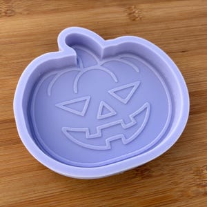 May include: A purple silicone mould shaped like a jack-o'-lantern. The mould has a detailed design of a pumpkin face with a wide smile, triangular eyes, and a nose.