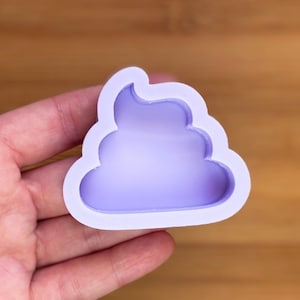 Puede incluir: Un molde de silicona de color púrpura claro con forma de pila de caca. El molde tiene un diseño suave y redondeado con un contorno detallado. El molde se sostiene en la mano de una persona contra un fondo de madera borroso.