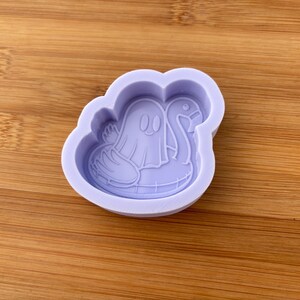 May include: A lavender silicone mold featuring a cartoon ghost floating on a swan-shaped pool float. The ghost has two oval eyes and a smiling mouth. The mold is on a wooden surface.