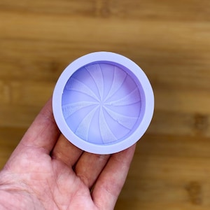 Puede incluir: Un molde redondo de silicona de color lavanda con un diseño en espiral. El molde se sostiene en una mano y está diseñado para manualidades o repostería. El patrón en espiral está en relieve, creando una superficie texturizada.