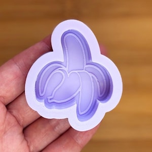Puede incluir: Un molde de silicona de color lila claro con forma de plátano pelado. El molde tiene un diseño detallado con múltiples capas, creando un efecto 3D. La forma de plátano se sostiene en una mano.