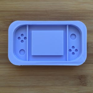 May include: A light purple silicone mould shaped like a handheld video game console. The mould has a rectangular screen area and two button areas with raised circles.