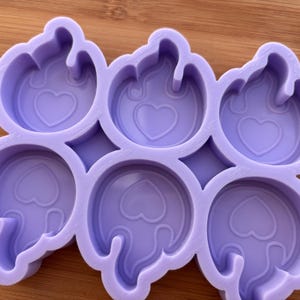 Può includere: Uno stampo in silicone lavanda con sei cavità circolari, ciascuna con un design a forma di cuore all'interno di un contorno a forma di fiamma. Lo stampo è progettato per la creazione di piccoli oggetti decorativi.