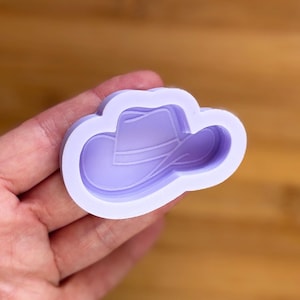 Puede incluir: Un molde de silicona de color lavanda con forma de sombrero de vaquero. El molde se sostiene en una mano y está diseñado con líneas detalladas para definir la forma del sombrero. El fondo es una superficie de madera borrosa.