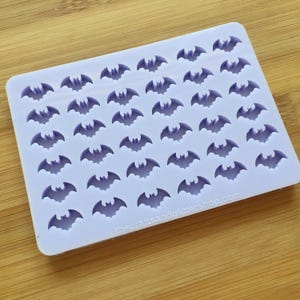 May include: A purple silicone mould with 42 bat-shaped cavities. The mould is on a wooden surface. The text "thecraftsandglittershop.com" is printed on the mould.