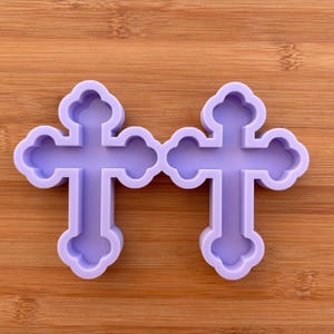Può includere: Due stampi a forma di croce viola chiaro con un design decorativo e arrotondato. Gli stampi sono realizzati in un materiale flessibile e sono appoggiati su uno sfondo di legno. Le croci sono probabilmente per l'artigianato o la cottura.