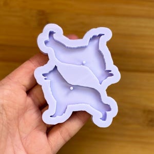 Pode incluir: Um molde de silicone roxo claro com duas silhuetas de cães. O molde é segurado em uma mão e é projetado para artesanato. As formas dos cães são detalhadas e prontas para resina ou outros materiais.