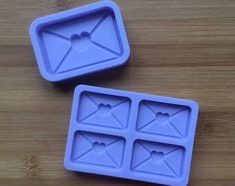 May include: Two light purple silicone molds for crafting. One mold is a single rectangular unit featuring a recessed envelope design with a heart-shaped seal. The second mold is a larger rectangular tray containing four identical envelope cavities.