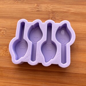 Può includere: Uno stampo in silicone lilla chiaro con quattro forme di torce stilizzate, ciascuna con un dettaglio a cuore. Lo stampo è progettato per l'artigianato e si trova su una superficie di legno.