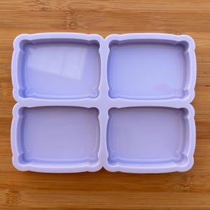 Può includere: Uno stampo in silicone lilla chiaro con quattro cavità rettangolari a forma di cuscino. Lo stampo è progettato per la creazione di piccoli oggetti decorativi. Lo stampo è su una superficie di legno.