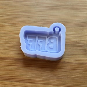 Peut inclure: Un moule en silicone violet clair avec les lettres "BFF" en police de caractères. Le moule a une boucle en haut pour l'attacher à un porte-clés ou autre. Le moule est sur une surface en bois.