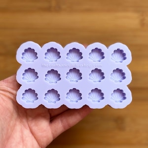Può includere: Uno stampo in silicone viola chiaro con quindici cavità a forma di conchiglia. Lo stampo ha bordi arrotondati e il testo "thecraftsandglittershop.com" è stampato in alto. Lo stampo è tenuto in mano su uno sfondo di legno.