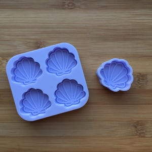 Pode incluir: Um molde de silicone roxo com quatro formas de conchas. Há também um molde de concha individual ao lado do molde maior.