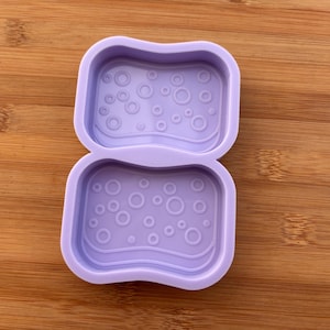 Può includere: Uno stampo in silicone viola chiaro con forma rettangolare e bordi arrotondati. Lo stampo è diviso in due sezioni, ciascuna con un design ondulato e impronte circolari di varie dimensioni, perfetto per sapone o resina.