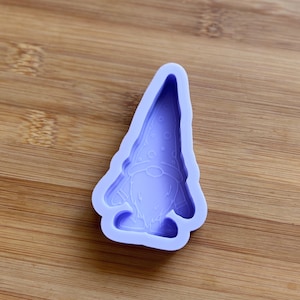 Puede incluir: Un molde de silicona lavanda con forma de gnomo. El gnomo tiene un gorro puntiagudo con lunares, una larga barba y una cara sonriente. El molde está diseñado para manualidades y la creación de artículos decorativos.