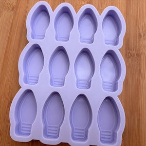 Può includere: Uno stampo in silicone viola chiaro con dodici cavità a forma di lampadine natalizie. Lo stampo è progettato per l'artigianato o la cottura al forno, con ogni cavità della lampadina con una base dettagliata. Lo sfondo è una superficie di legno.