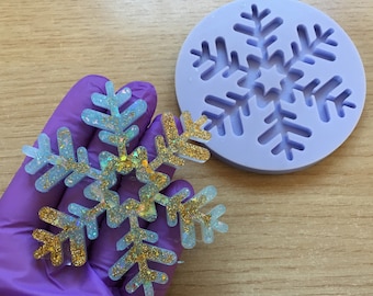 Op de afbeelding: Een sneeuwvlokvormig harsgietstuk met goudkleurige en iriserende glitters naast een ronde, lichtpaarse siliconen mal. De afgewerkte sneeuwvlok heeft een doorschijnende blauwe basis met gouden glitteraccenten in de structuur.