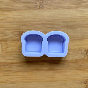 Puede incluir: Un molde de silicona morado con dos cavidades en forma de pan. El molde está diseñado para hacer pequeños objetos decorativos con forma de pan.